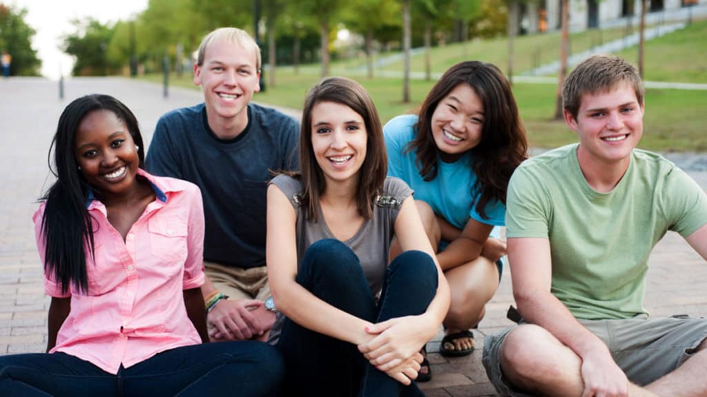 Diverse group of five young adults sitting outside on pavement and smiling